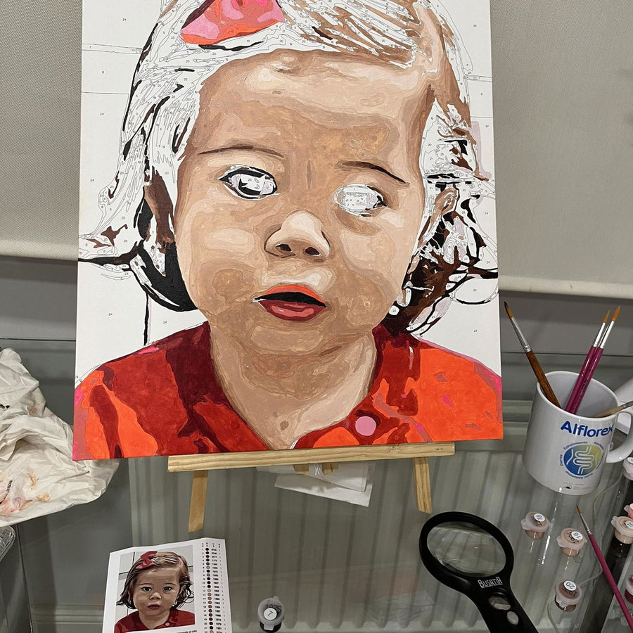 Retrato de una niña a medio pintar en el caballete, con la foto de referencia al lado